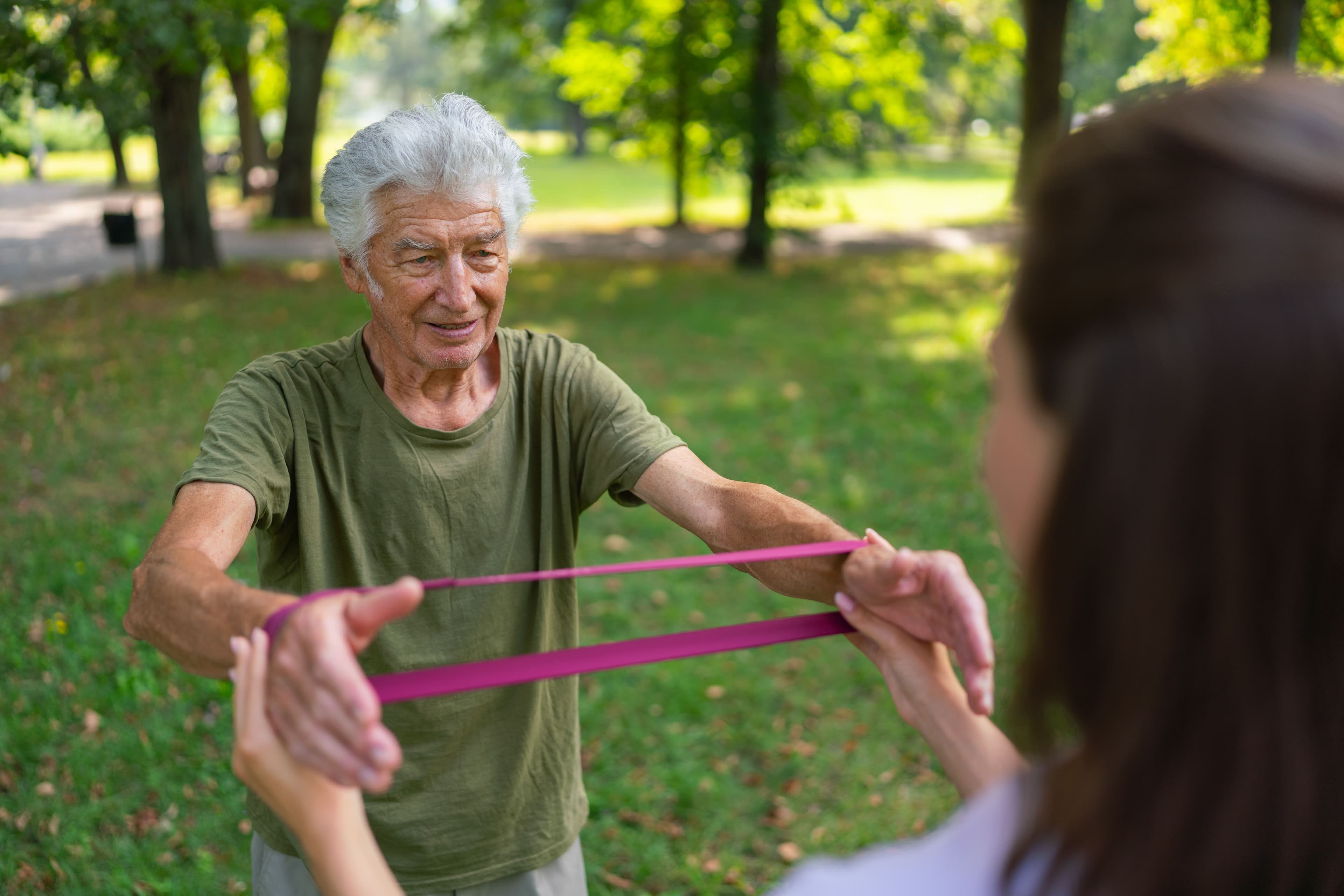 an old man in a park stretching an exercise resistance band between his wrists under the guidance of a female Physiotherapist