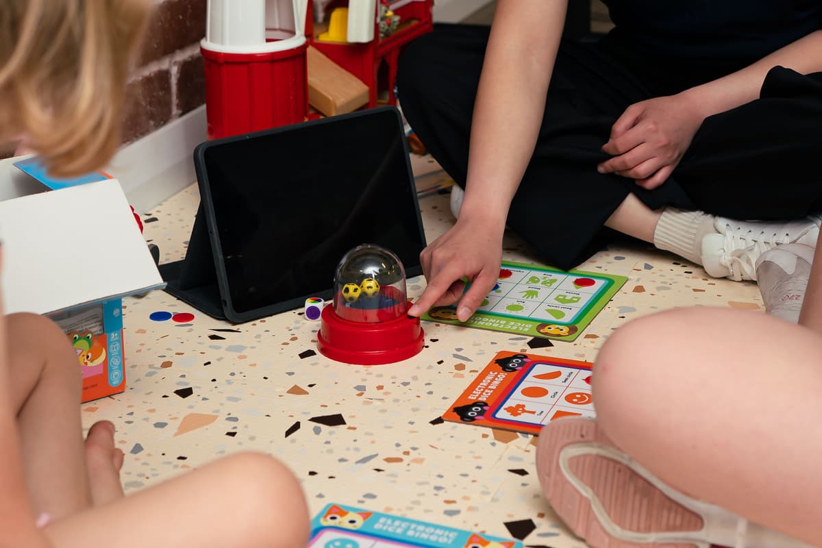 Two children and an adult sitting down on a play mat, and playing a game with dice in a plastic sphere