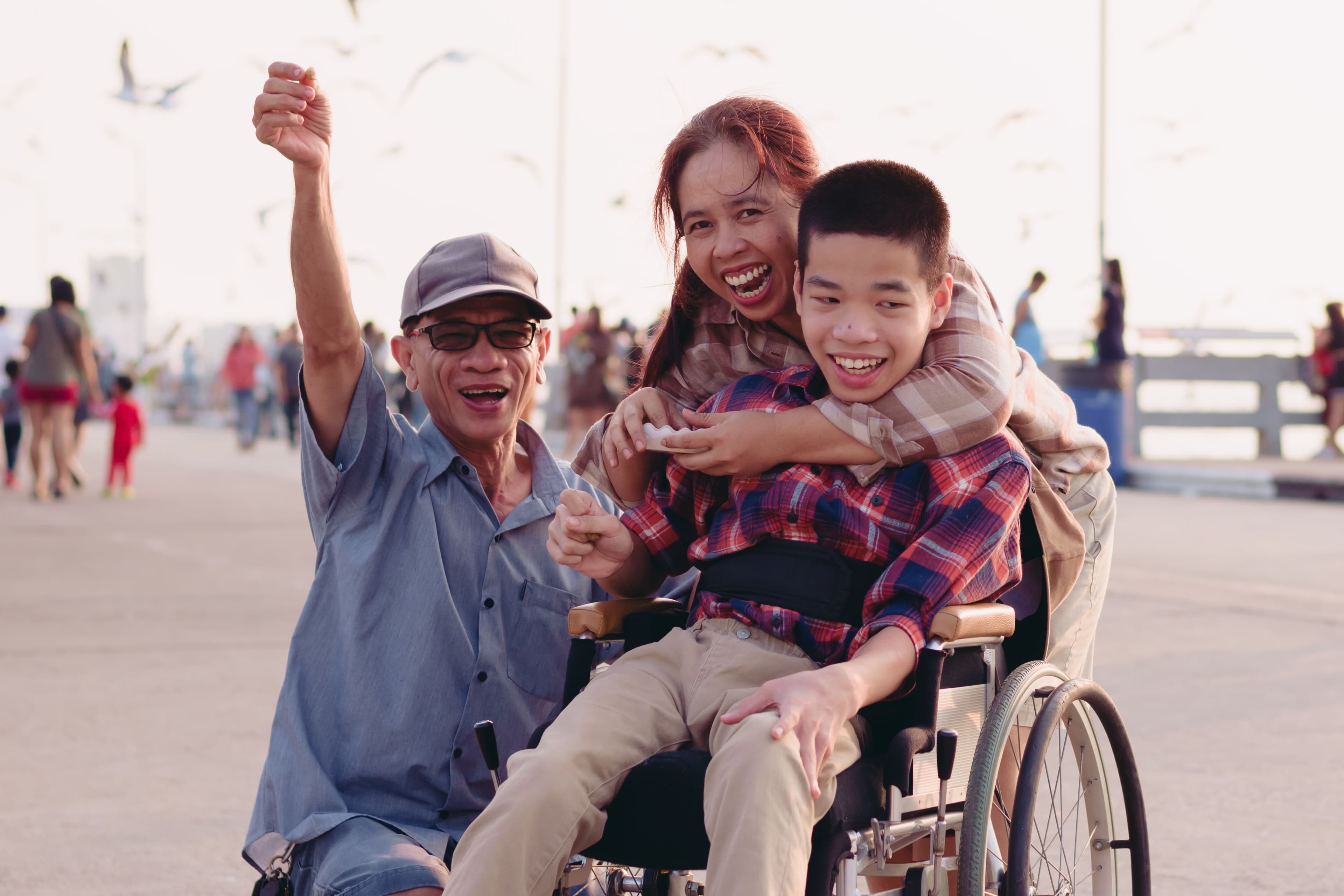 young smiling boy in wheelchair being hugged from behind by smiling woman with smiling man crouching alongside