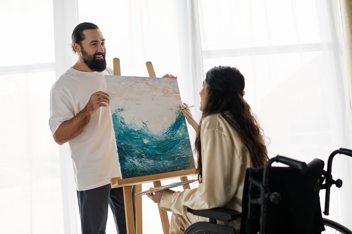 A lady in a wheelchair painting a beautiful portrait while a man holds the canvas