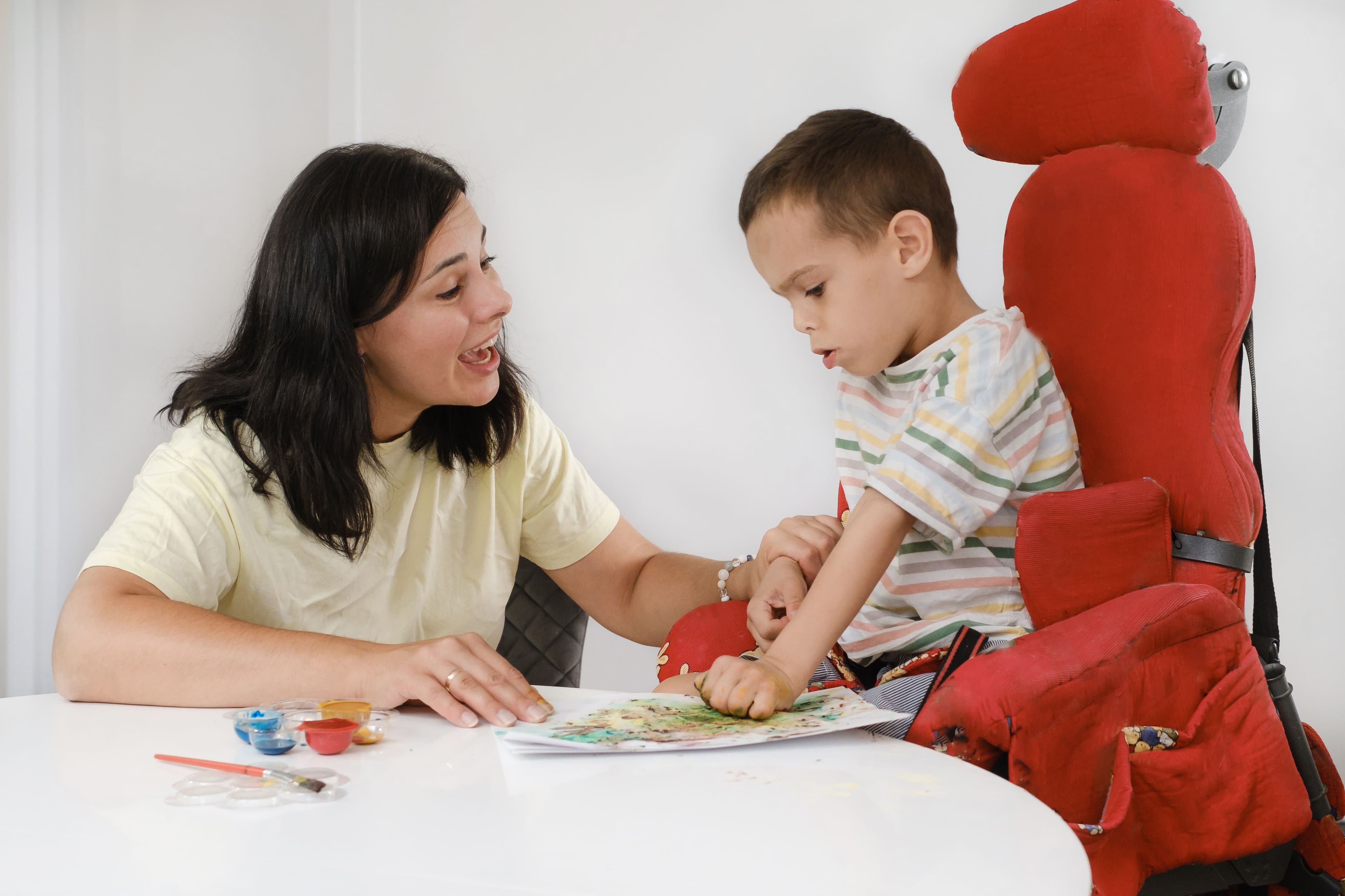 a woman sitting at a table and kindly engaging with a young boy with a physical disability who sitting in special booster chair with a drawing in front of him